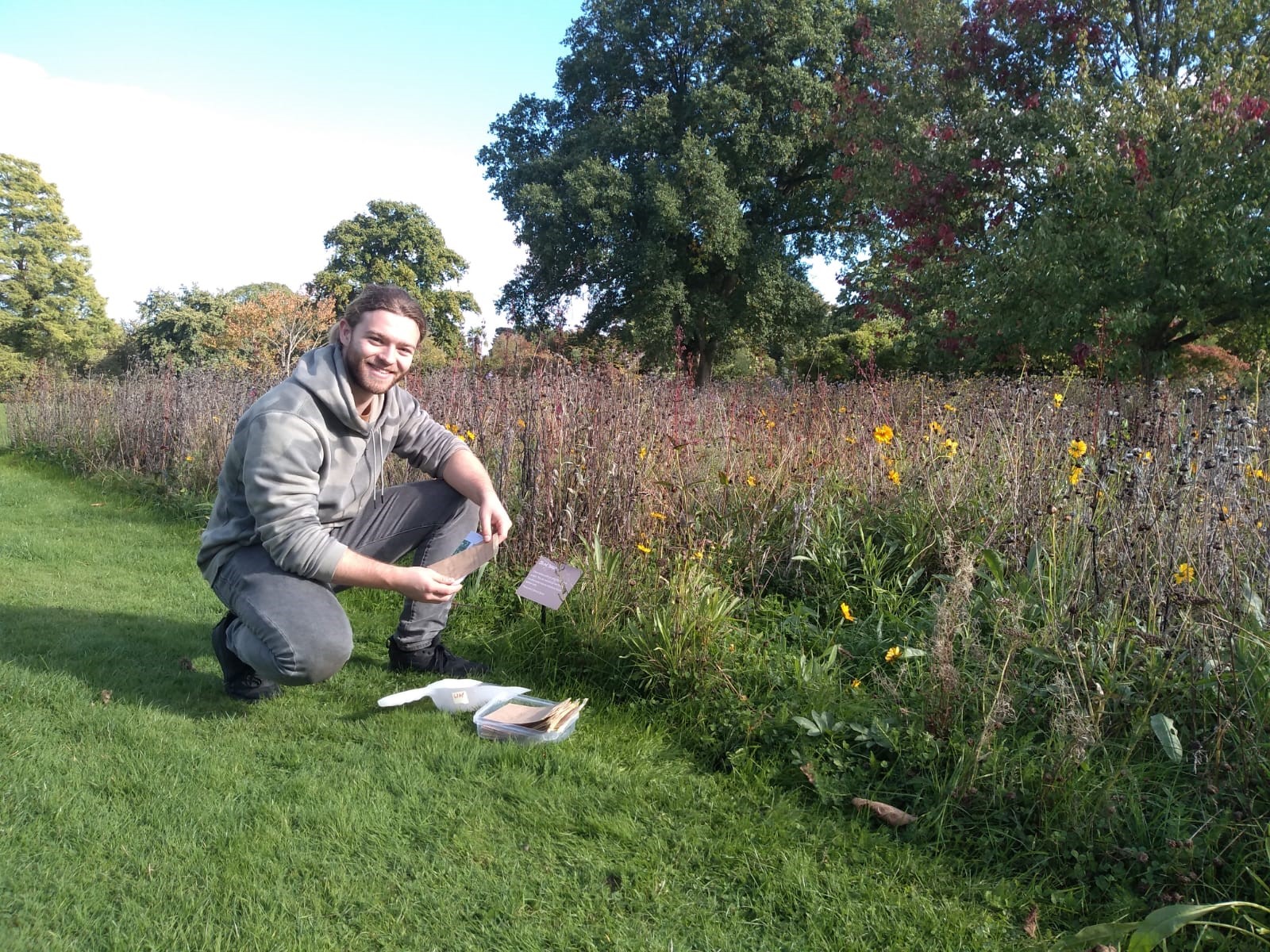 A man knelt down on a moxed piece of grass next to aa taller sward of plants some with yellow flowers. He is holding a paper envelope.
